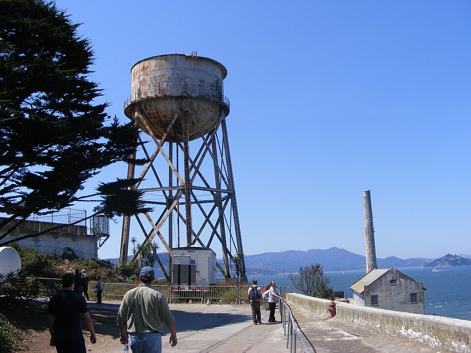 The Water Tower on Alcatraz, with visitors walking the path beside it and the Marin hills across the bay