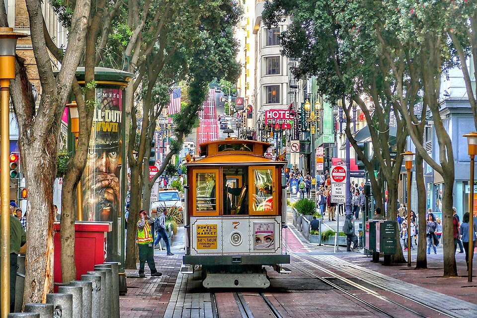 A cable car coming down the tree-lined lower end of Powell Street, flower stands on the sidewalk