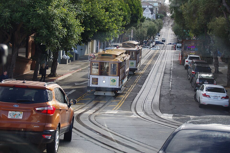 A cable car climbing a steep street between parked cars