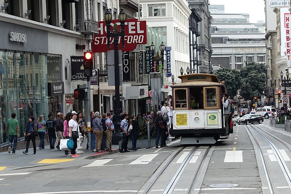 Riders crowding the curb as a cable car stops on Powell Street by the Hotel Stratford