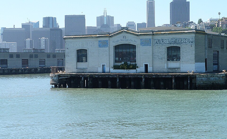 A pier shed standing on its piles over the bay, downtown towers behind it