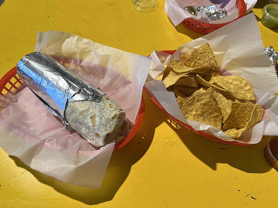 A Mission burrito wrapped in foil, with a basket of chips