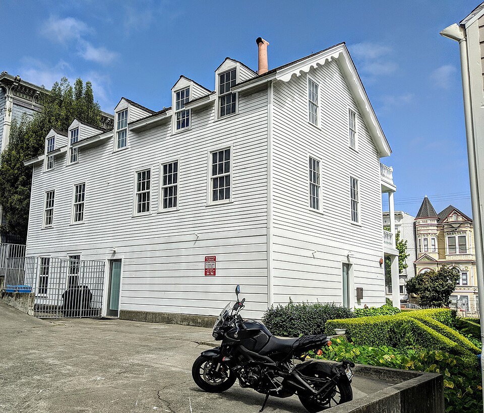 A plain white clapboard house with dormer windows, among its Victorian neighbors