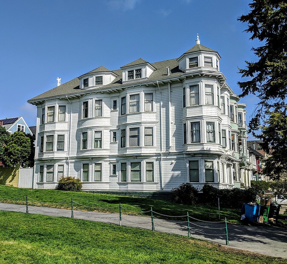 A white turreted Victorian apartment house above a green lawn